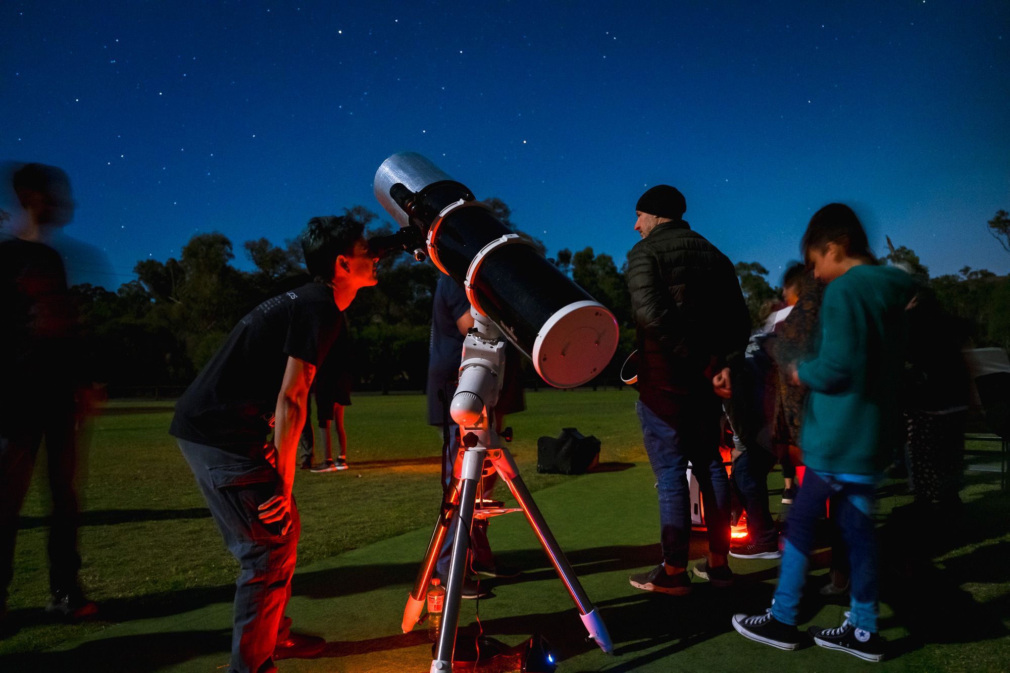 A group of people gather around a large telescope on a tripod, looking up at a starry night sky with trees in the background.