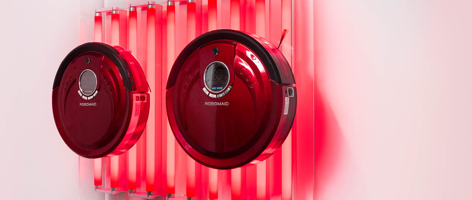 Two red robotic vacuum cleaners are mounted on a white wall with vertical red lights behind them.