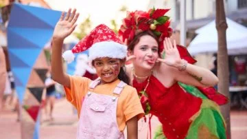 Smiling girl in Santa hat with face paint waves beside a performer in a festive red and green costume.