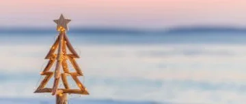 Wooden Christmas tree with fairy lights glowing against a soft pastel beach sunset.