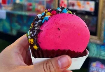 Bright pink ice cream sandwich topped with colorful candy sprinkles and cookie crumbs in a paper holder.