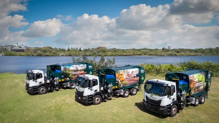 Three waste collection trucks parked on grass beside a river, promoting recycling and waste services under a blue sky.