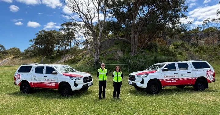 bushfire safety officers standing between two marked Bushfire Safety vehicles in a grassy reserve.