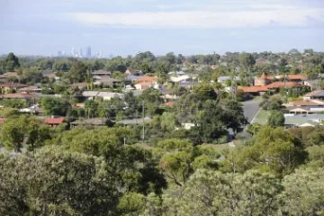 Residential suburb surrounded by trees, with streets and rooftops visible and a distant city skyline on the horizon.