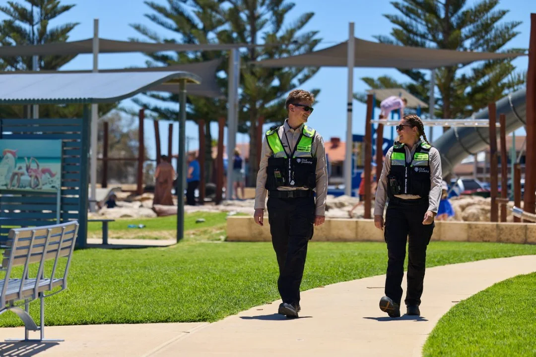 Two Community safety officers walking along a path through a park playground on a sunny day.