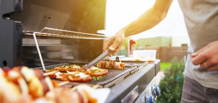 Person cooking sausages and skewers on an outdoor barbecue using tongs in a backyard setting.