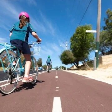 Child wearing helmet rides bicycle on shared path, following another cyclist on a sunny day.