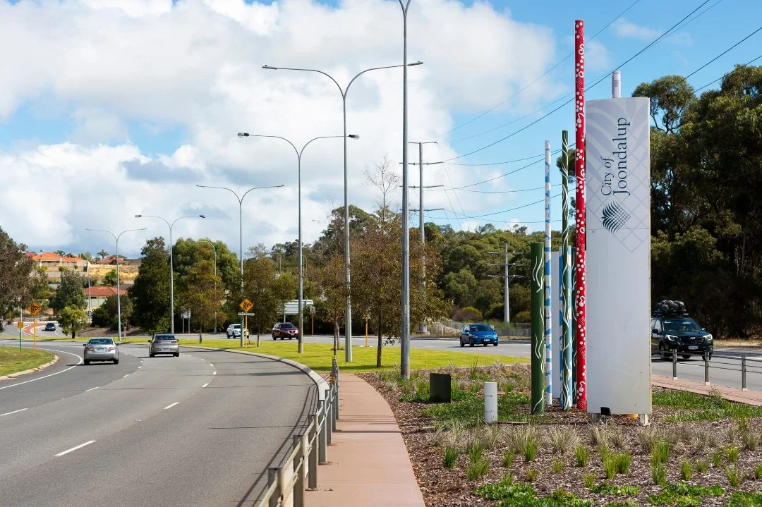 Roadside entrance sign for the City of Joondalup beside a multi-lane road with cars, streetlights and landscaped verge.
