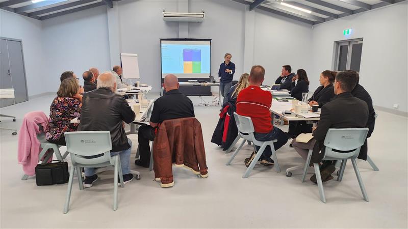 Group of adults seated in a meeting room, listening to a presenter beside a screen displaying a colourful chart