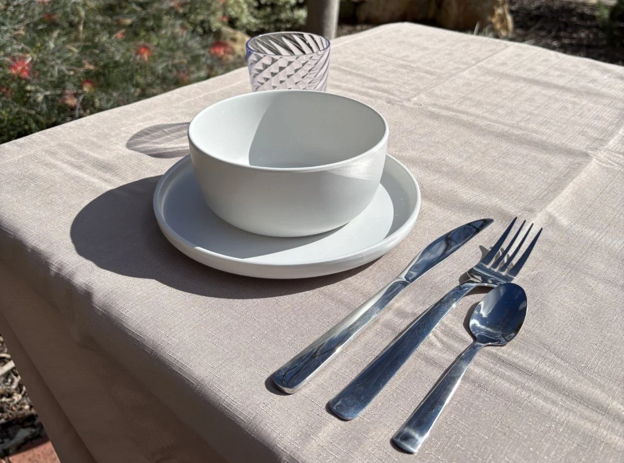 Outdoor table setting with bowl, plate, glass, knife, fork, and spoon on a beige tablecloth.