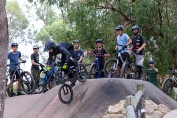 A group of kids on bikes at a dirt pump track. One rider in black gear is going over a bump while others watch.