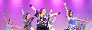 Group of performers in colorful outfits dancing on stage under pink and purple lighting with arms raised. 