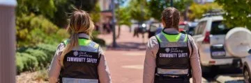 Two community safety officers in high-visibility vests walking along a pedestrian street.