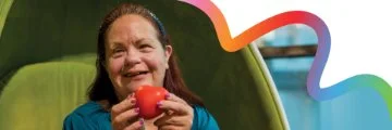 Lady holding a miniature heart in her hands with a welcoming smile, on a green background including a rainbow wave edge.