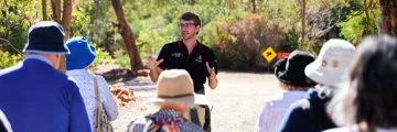 Man in black polo speaks to group wearing sun hats on bush trail during outdoor guided talk.