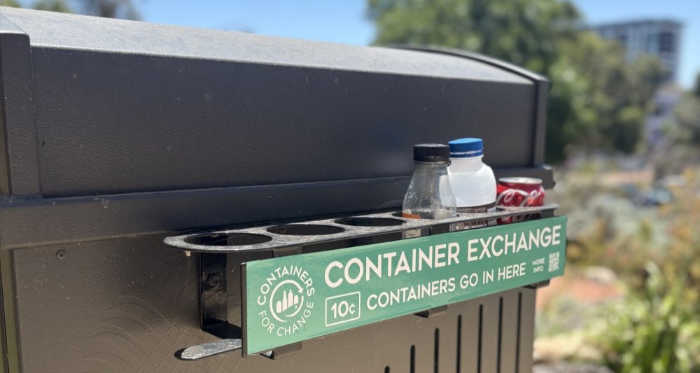 Outdoor bin with a “Containers for Change” container exchange slot holding bottles and cans against a park backdrop