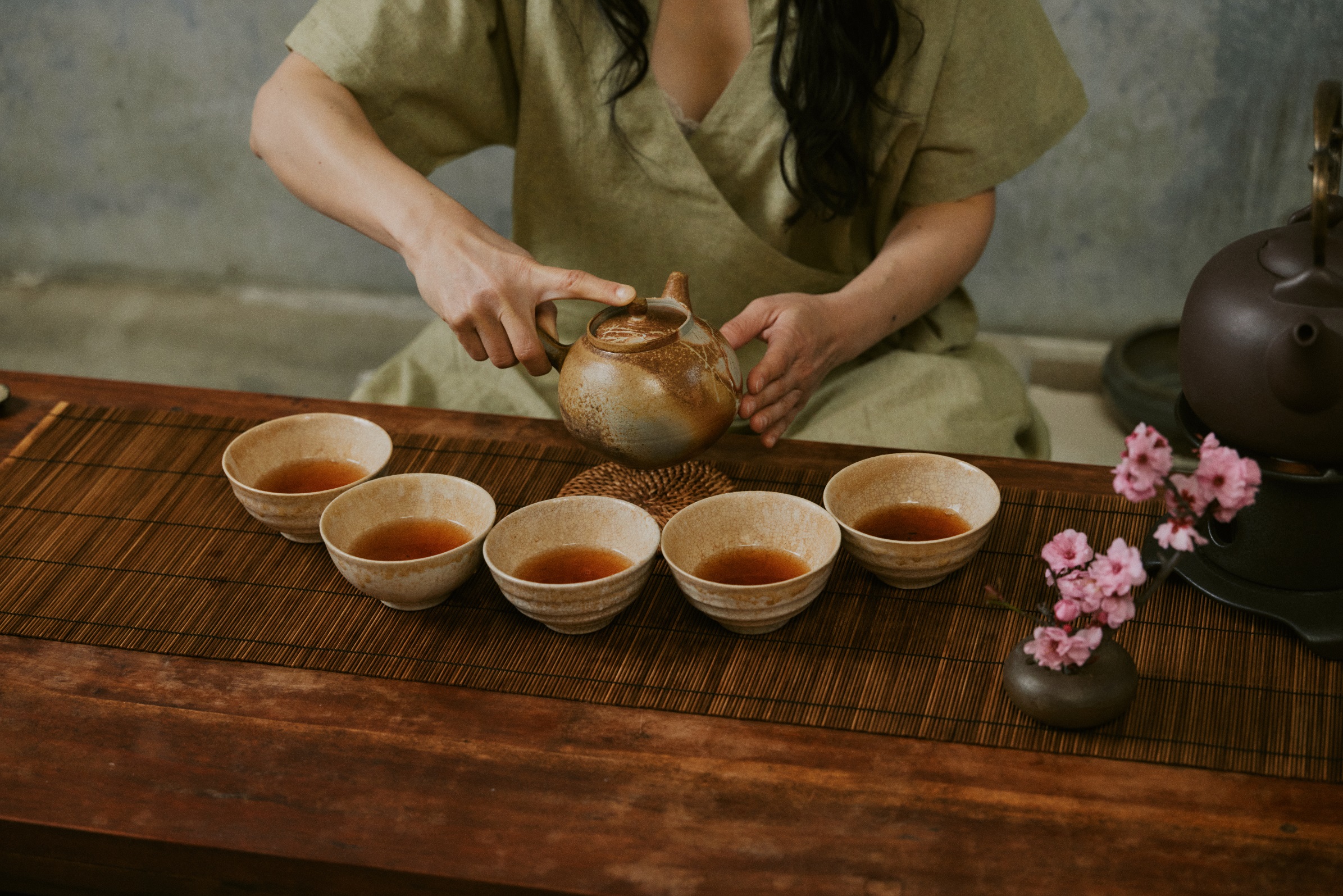 A person holds a ceramic teapot surrounded by small bowls, arranged on a bamboo mat on a wooden table.